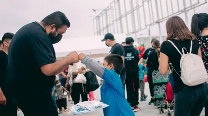 Man and boy wearing masks give a fist bump outdoors at a crowded event or market with people walking around.