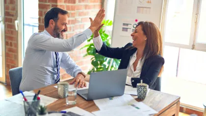Two coworkers smiling and giving a high-five over a desk with laptop and documents in bright office.