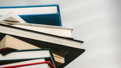 Stack of various hardcover books piled up with a plain gray background, viewed from a low angle.