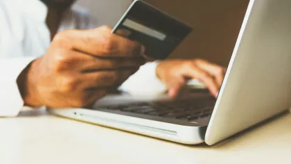 Person holding credit card while using laptop for online payment on desk.