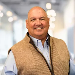 Smiling bald man wearing a light blue shirt and beige zip-up sweater vest in a bright indoor setting